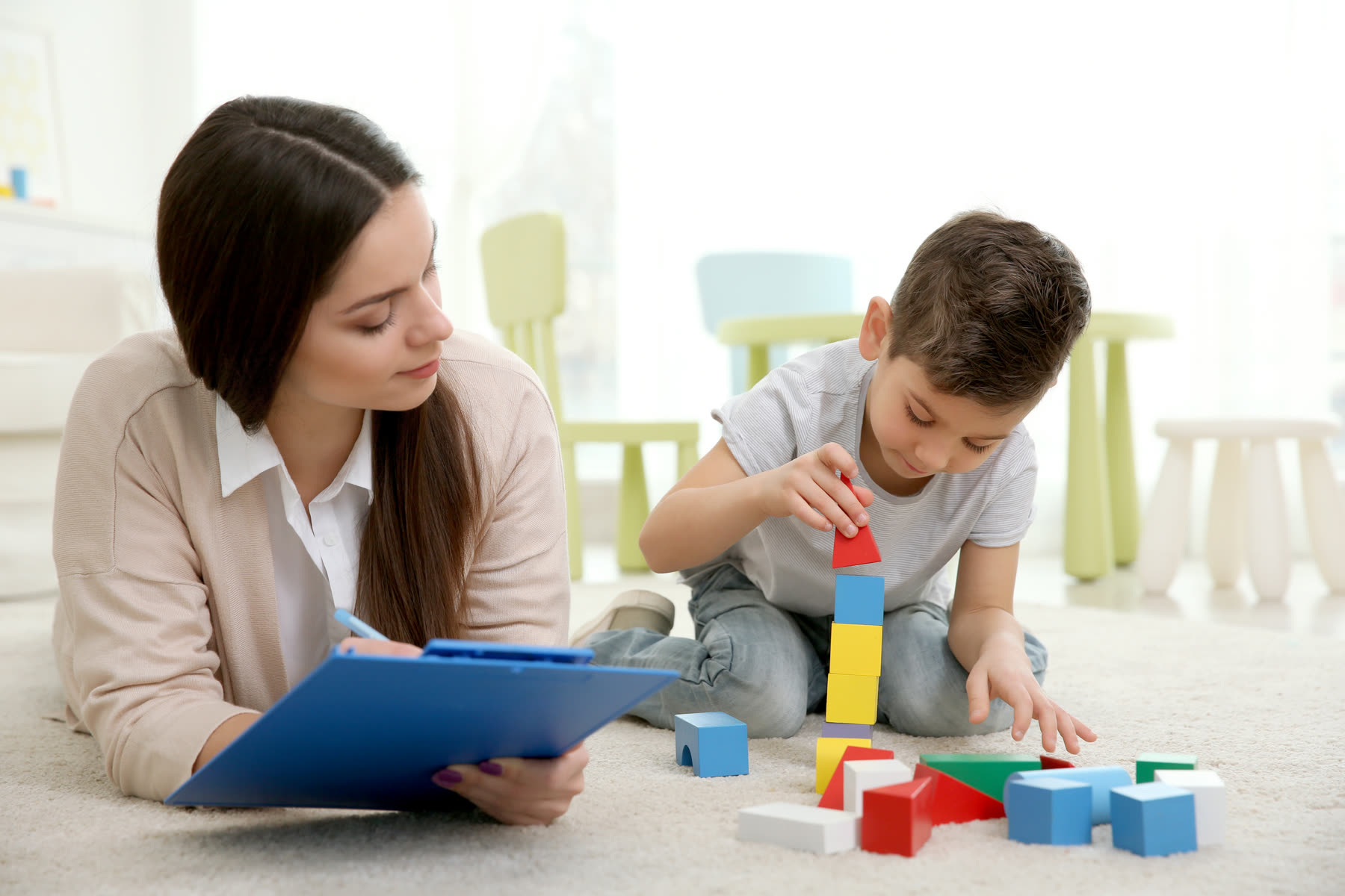A clinician with a clipboard watching a young boy build with blocks
